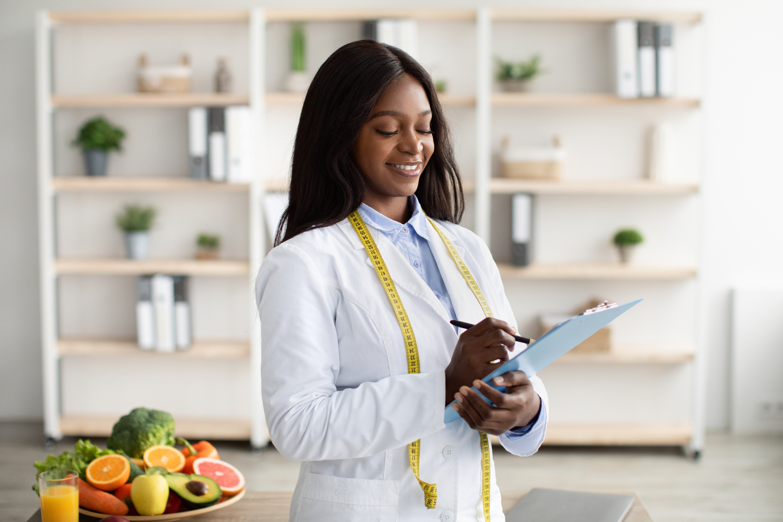 Cheerful black nutritionist with clipboard prescribing healthy diet or creating plant based meal plan for patient at weight loss clinic, copy space