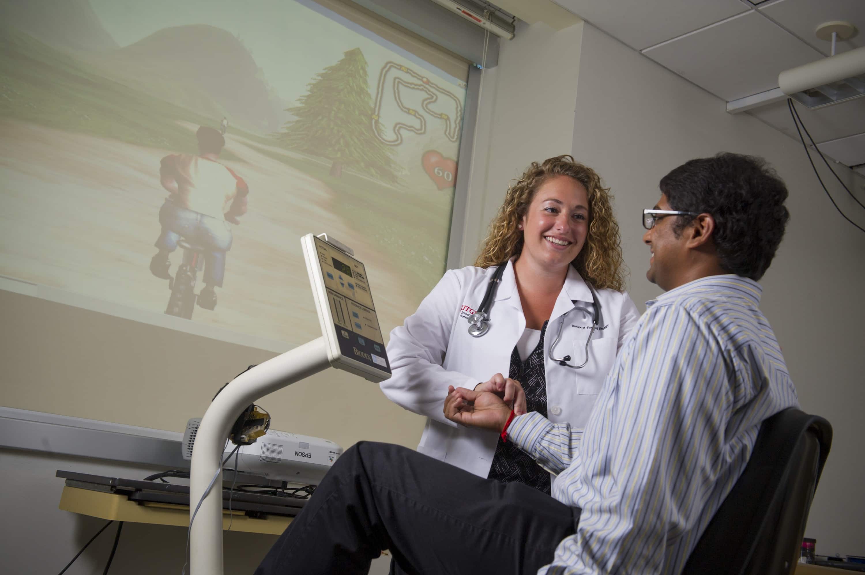 Rivers Lab student researcher monitors heart rate of study participant on stationary bike with video game image projected on the wall.